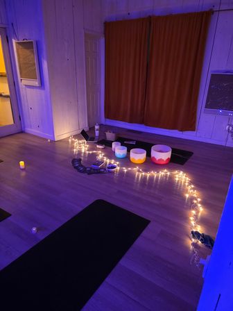 Cozy meditation studio sound-bath setup with yoga mats, three colorful crystal singing bowls, string fairy lights and LED candles under soft purple LED lighting and closed curtains