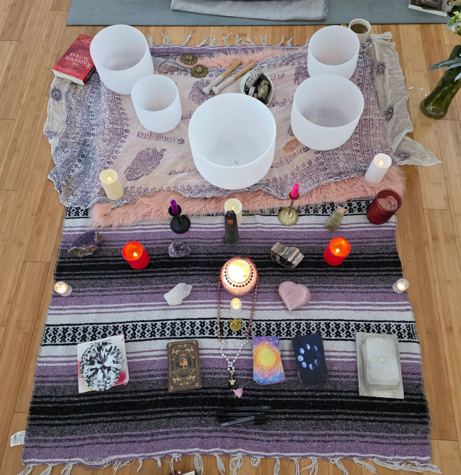 Top-down view of a boho home meditation altar on hardwood floor: white crystal singing bowls, lit candles, assorted crystals including a heart-shaped rose quartz, tarot and oracle decks, mala beads and ritual tools arranged on layered purple and pink blankets.