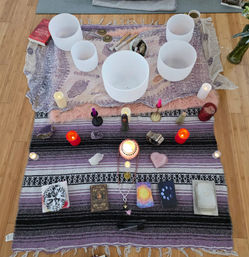 Top-down view of a boho home meditation altar on hardwood floor: white crystal singing bowls, lit candles, assorted crystals including a heart-shaped rose quartz, tarot and oracle decks, mala beads and ritual tools arranged on layered purple and pink blankets.