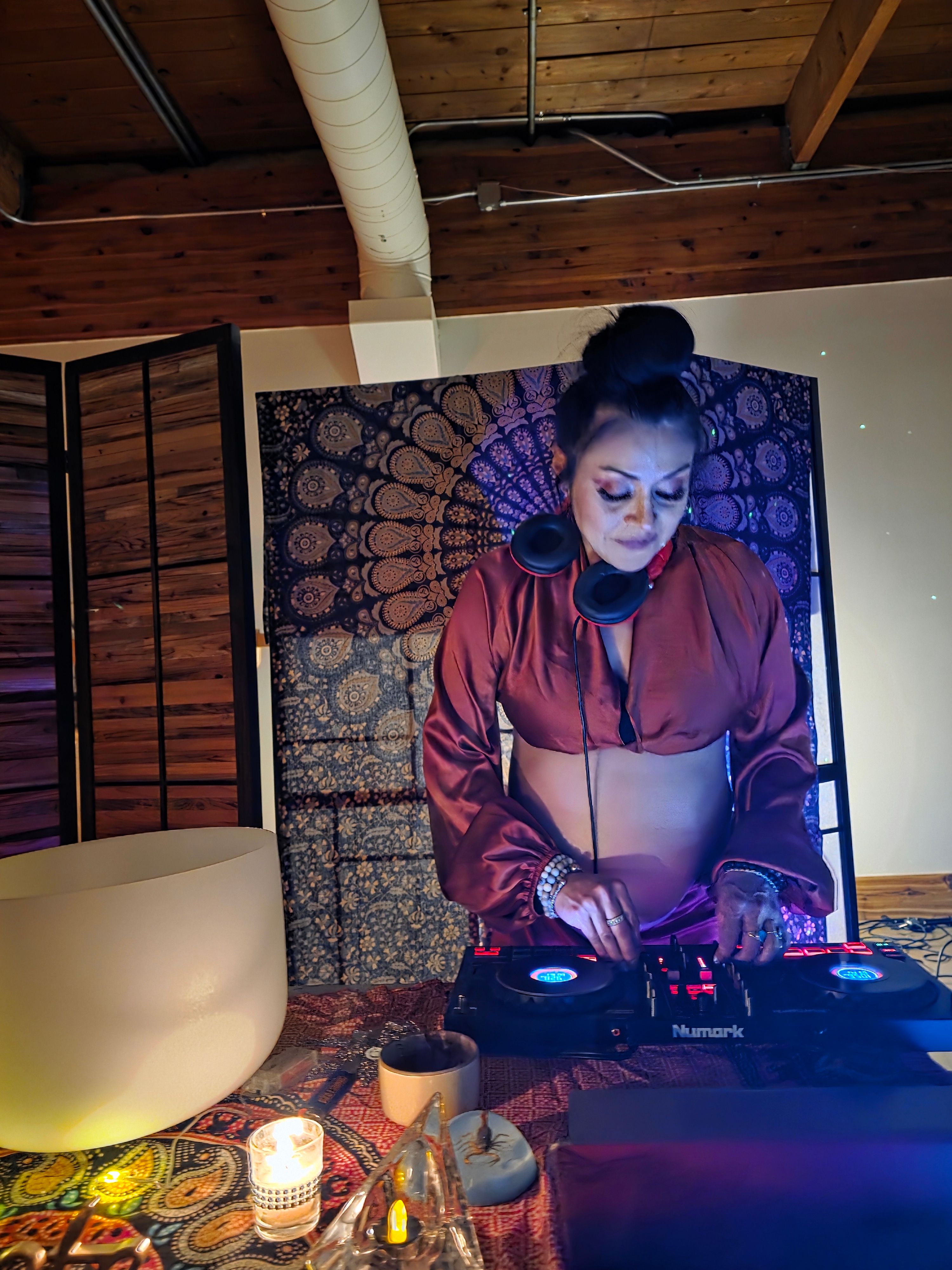 Boho DJ in a cozy loft studio, headphones around her neck as she mixes on Numark controllers in front of a mandala tapestry, with candles, a crystal singing bowl and patterned tablecloth creating warm ambient vibes.