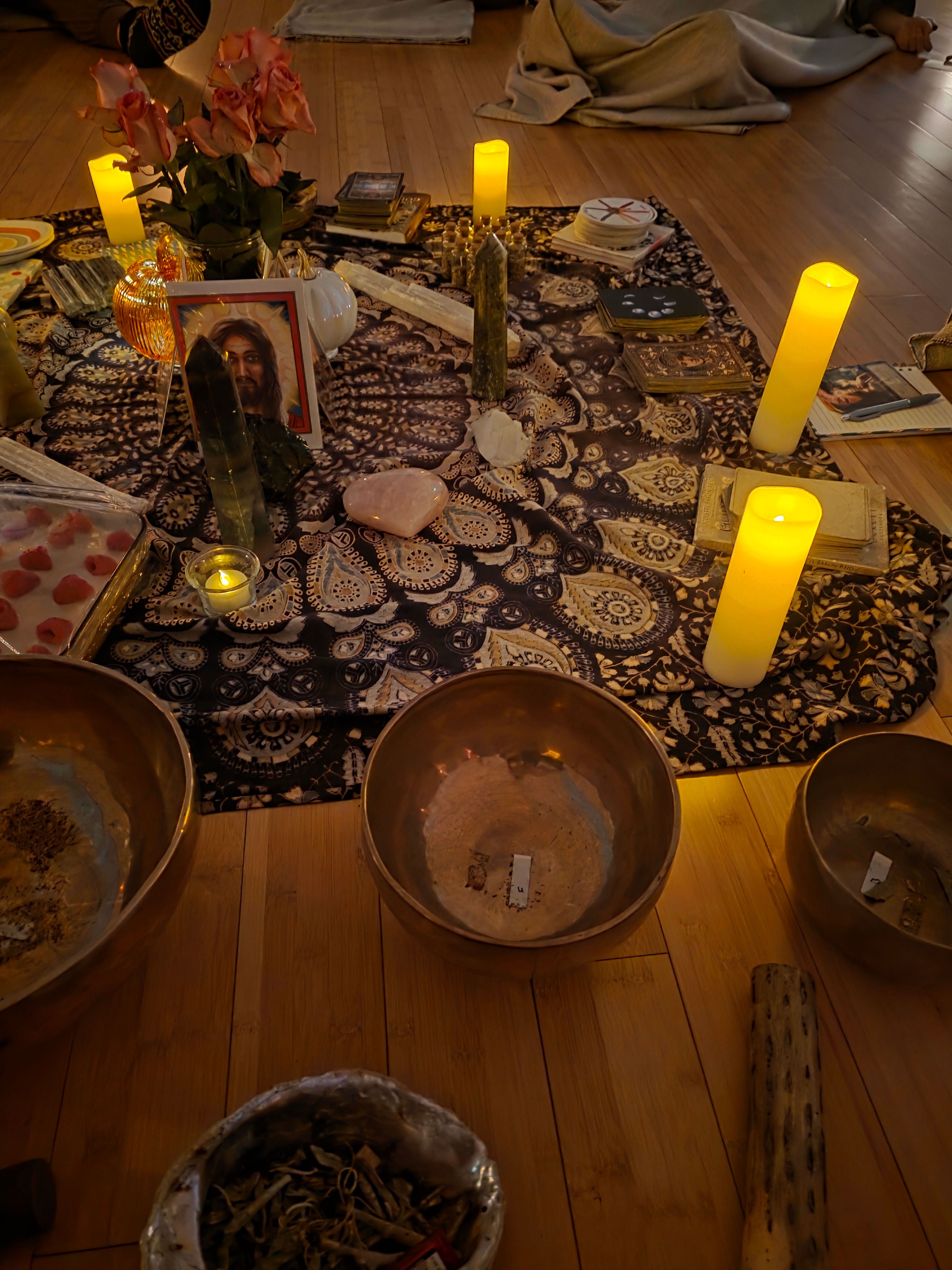 Cozy indoor meditation altar on hardwood floor with patterned cloth, glowing pillar candles, singing bowls, crystals, roses, oracle cards and a framed portrait for a healing circle.
