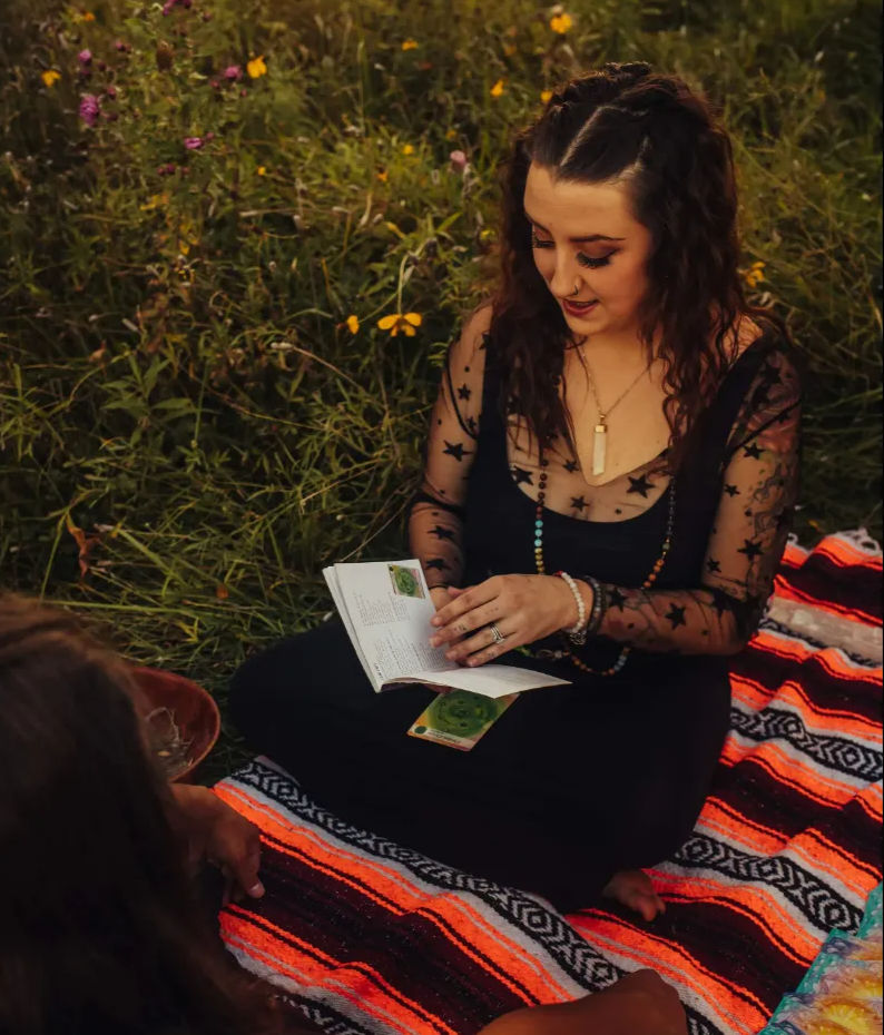 Woman in star-print sheer top seated on a bright striped blanket in a wildflower meadow, holding a booklet and reading tarot cards in a relaxed bohemian outdoor scene.