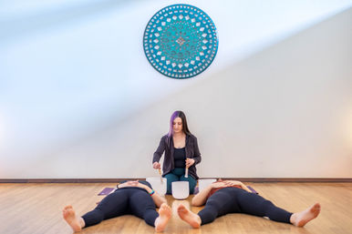 Instructor leading a sound bath in a bright yoga studio, seated on the floor between crystal singing bowls while two participants lie in savasana on mats under a turquoise mandala wall art — meditation, relaxation, and wellness session.