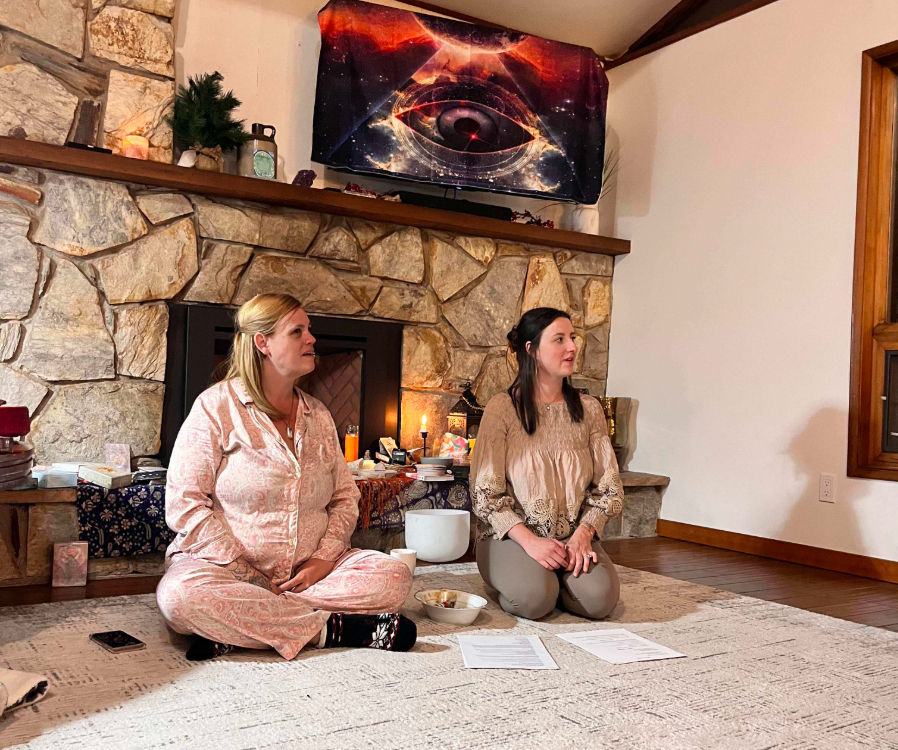Two women seated on a rug in a cozy home gathering in front of a stone fireplace with candles, crystals and a singing bowl, cosmic eye tapestry hanging above.