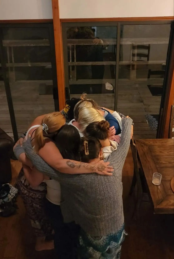 Six people sharing a close group hug in a cozy living room with wooden floors and sliding glass doors — warm evening gathering.