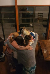 Six people sharing a close group hug in a cozy living room with wooden floors and sliding glass doors — warm evening gathering.