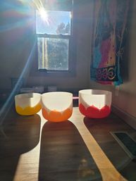 Sunlit meditation space with sunbeams from a window illuminating three frosted crystal singing bowls in yellow, orange, and red on a wooden floor next to a colorful bohemian tapestry — sound healing setup.