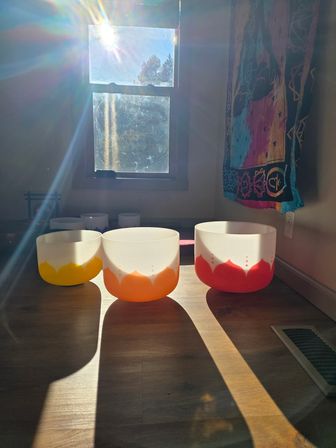 Sunlit meditation space with sunbeams from a window illuminating three frosted crystal singing bowls in yellow, orange, and red on a wooden floor next to a colorful bohemian tapestry — sound healing setup.