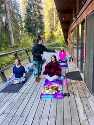Women taking a guided outdoor meditation on a rustic wooden cabin deck surrounded by tall evergreen trees, colorful yoga mats and cozy blankets for a peaceful forest retreat vibe.
