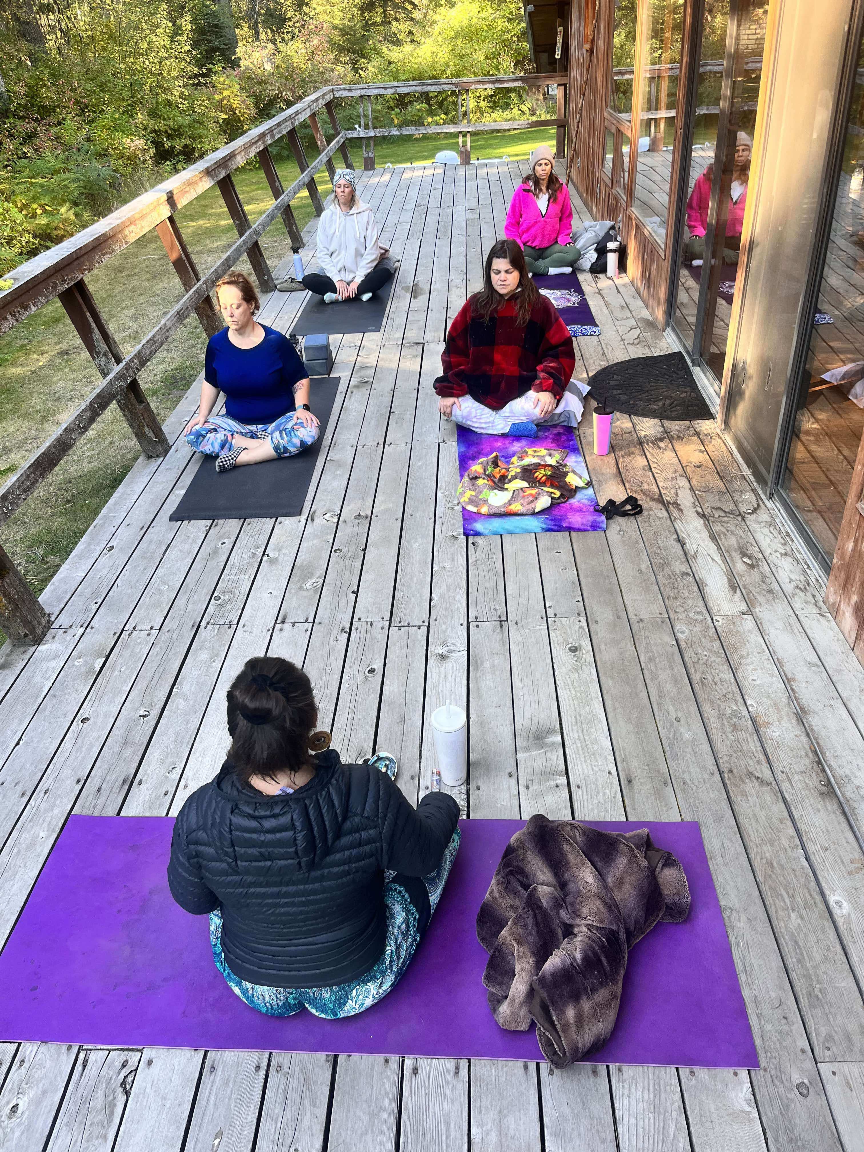 Outdoor yoga and meditation class on a weathered wooden deck by a glass-walled cabin — five people seated on colorful mats in meditative pose surrounded by green forest, retreat-style setting.