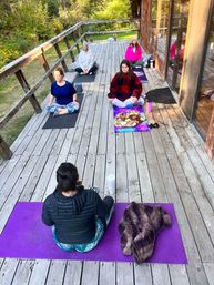 Outdoor yoga and meditation class on a weathered wooden deck by a glass-walled cabin — five people seated on colorful mats in meditative pose surrounded by green forest, retreat-style setting.