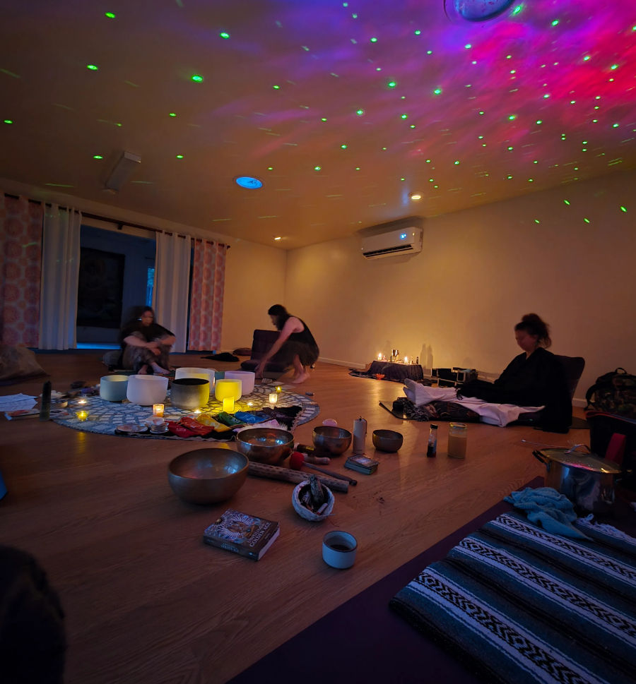 Dimly lit wellness studio with green and purple starry ceiling lights, participants seated around a circle of singing bowls, candles, crystals and mats for a relaxing group sound‑bath meditation