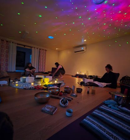 Dimly lit wellness studio with green and purple starry ceiling lights, participants seated around a circle of singing bowls, candles, crystals and mats for a relaxing group sound‑bath meditation
