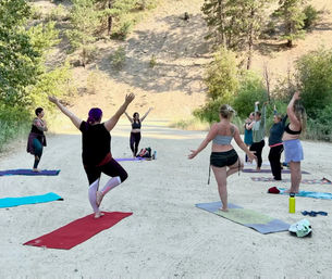 Outdoor group yoga class on a sunlit dirt road in a forested hillside, women on colorful mats practicing balancing poses with arms raised.