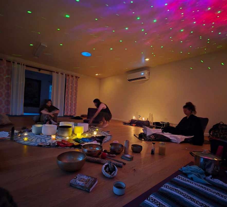 Cozy dim wellness studio with three people seated on mats around crystal singing bowls, candles, and small instruments; green and pink laser lights speckle the ceiling.