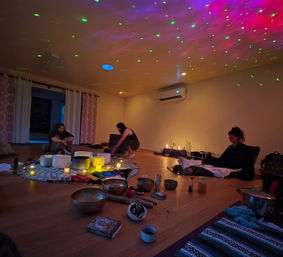 Cozy dim wellness studio with three people seated on mats around crystal singing bowls, candles, and small instruments; green and pink laser lights speckle the ceiling.