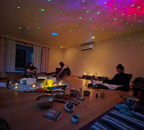 Cozy dim wellness studio with three people seated on mats around crystal singing bowls, candles, and small instruments; green and pink laser lights speckle the ceiling.
