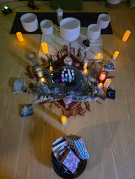 Top-down view of a cozy meditation altar with crystal singing bowls on a mat, glowing candles, crystals and sage, tarot/oracle cards, small paint bottles and a basket of notebooks.