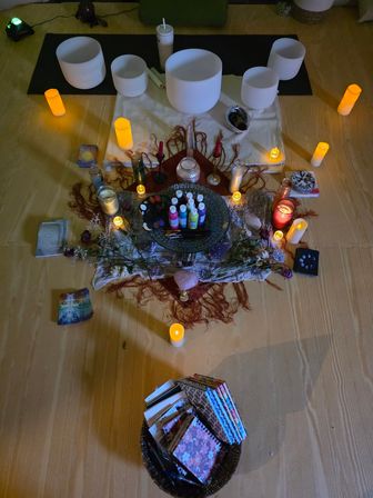 Top-down view of a cozy meditation altar with crystal singing bowls on a mat, glowing candles, crystals and sage, tarot/oracle cards, small paint bottles and a basket of notebooks.