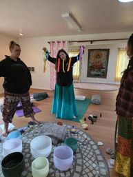 Group sound-healing workshop in a sunlit meditation studio: three women on wooden floors, one holding colorful ribbons, crystal singing bowls and cushions on a patterned rug, yoga mats and a Buddha tapestry in the background.