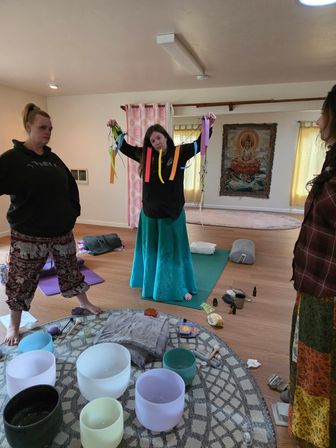 Group sound-healing workshop in a sunlit meditation studio: three women on wooden floors, one holding colorful ribbons, crystal singing bowls and cushions on a patterned rug, yoga mats and a Buddha tapestry in the background.