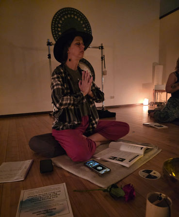 Person meditating cross-legged on a cushion in a dim wellness studio, hands in prayer, wearing a wide-brim hat, gong and salt lamp in the background, open workbook and phone on the mat and a rose on the wooden floor — yoga/meditation sound-bath session.