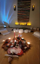 Cozy meditation/yoga studio set for a sound bath: floor cushions and crystal singing bowls surrounding a central altar with lit candles, crystals, tarot cards and red taper candles, woven wall hanging and blue star light projection