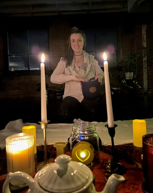 Smiling woman in a dim industrial loft seated behind a candlelit table with tall white candles, teapot, jar and heart-shaped LED light — cozy ritual scene.