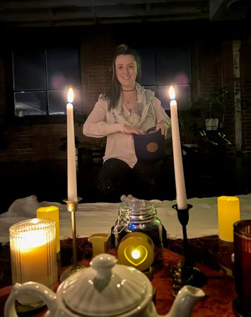 Smiling woman in a dim industrial loft seated behind a candlelit table with tall white candles, teapot, jar and heart-shaped LED light — cozy ritual scene.