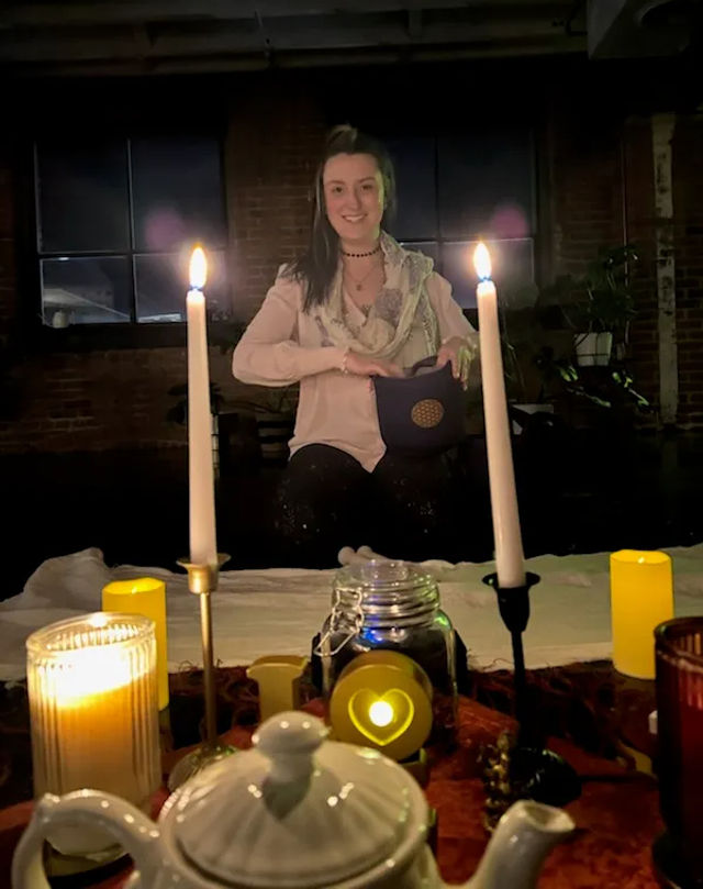 Smiling woman in a dim industrial loft seated behind a candlelit table with tall white candles, teapot, jar and heart-shaped LED light — cozy ritual scene.