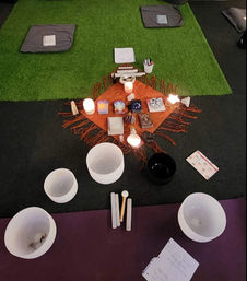Top-down view of a meditation altar in a studio: orange cloth on green turf with lit candles, crystals, oracle/tarot cards and small charms, surrounded by white and black singing bowls, mallets and floor cushions for a sound-healing circle.