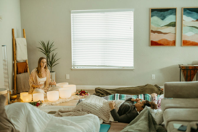 Person leads a relaxing sound-bath meditation in a cozy indoor studio: glowing crystal singing bowls on a rug, participants resting on blankets, a houseplant and towel ladder, and colorful abstract landscape prints on the wall.