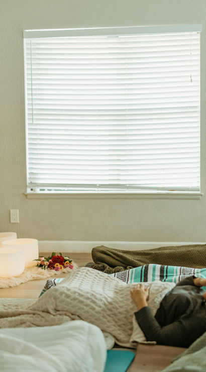 Person leads a relaxing sound-bath meditation in a cozy indoor studio: glowing crystal singing bowls on a rug, participants resting on blankets, a houseplant and towel ladder, and colorful abstract landscape prints on the wall.