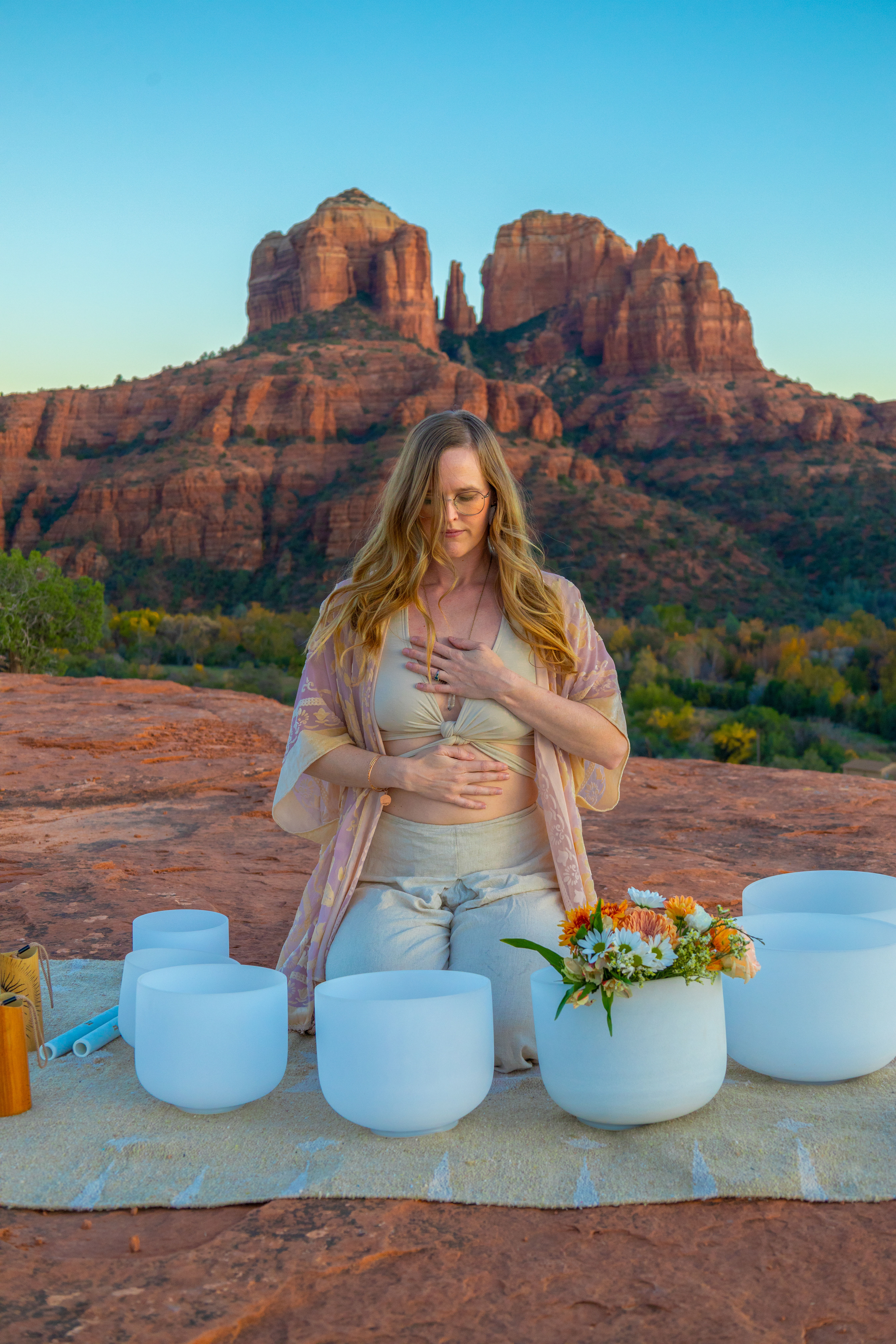 Woman meditating with hands on chest and abdomen amid white crystal singing bowls and flowers on a red-rock ledge with Cathedral Rock sandstone formations in Sedona, Arizona at golden hour.
