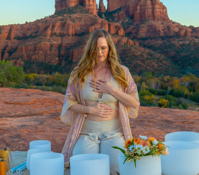 Woman meditating with hands on chest and abdomen amid white crystal singing bowls and flowers on a red-rock ledge with Cathedral Rock sandstone formations in Sedona, Arizona at golden hour.