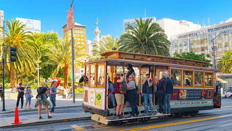 Historic San Francisco cable car filled with riders on a sunny day at Union Square, palm trees and downtown buildings in the background.