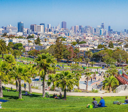 Sunny panoramic view of a busy urban park with palm trees and picnickers on grassy slopes, red pedestrian bridge and San Francisco skyline under a clear blue sky.