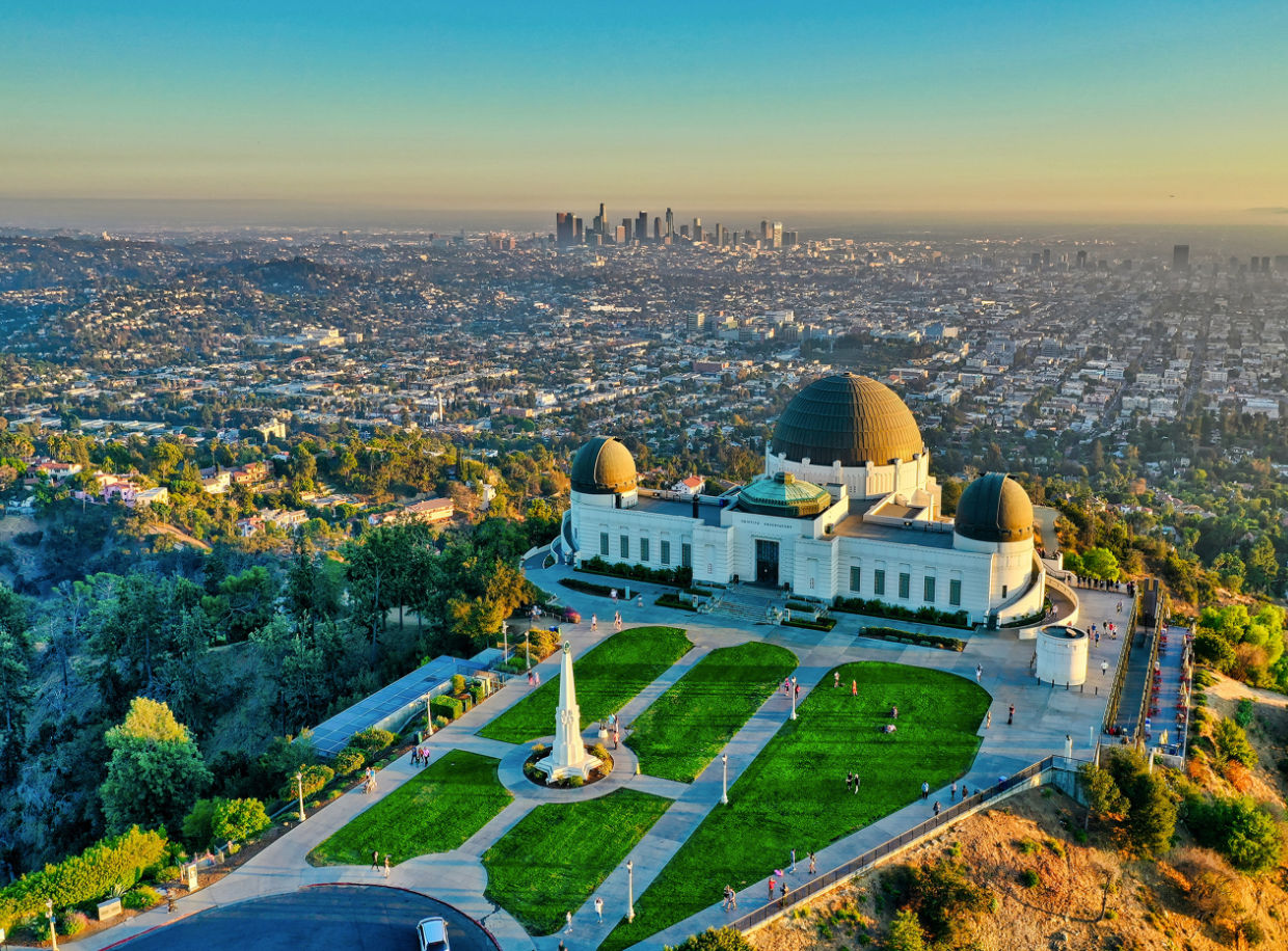 Aerial view of Griffith Observatory on a sunny Los Angeles hillside at golden hour, green lawns and walking paths in the foreground with the downtown LA skyline across the sprawling city
