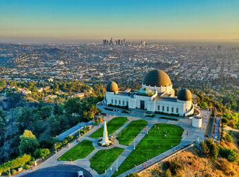 Aerial view of a domed hilltop observatory with green lawns and visitors, overlooking the Los Angeles skyline at golden hour