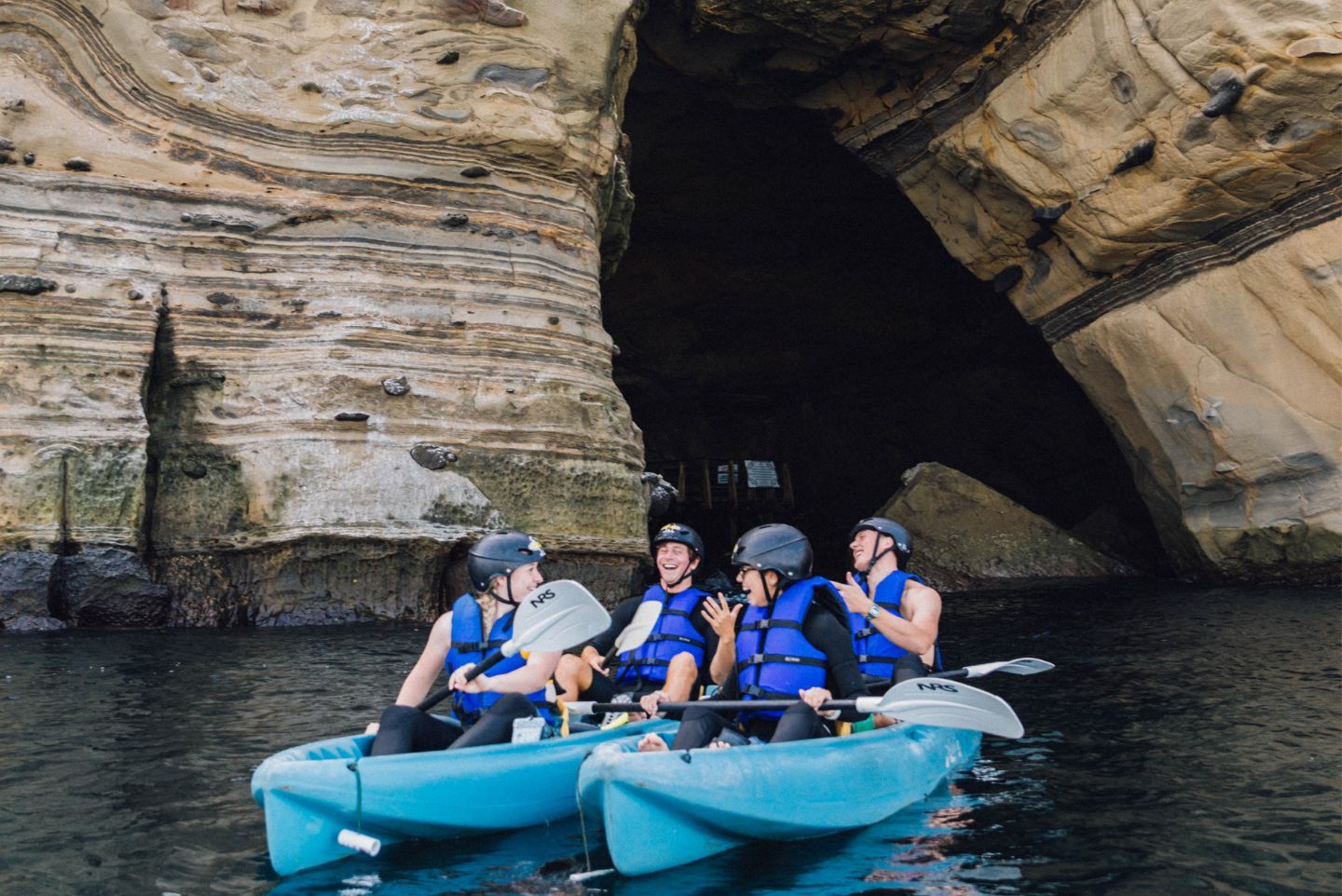 Four kayakers in blue life jackets and helmets laughing in two turquoise kayaks at the dark entrance of a sandstone sea cave along layered coastal cliffs