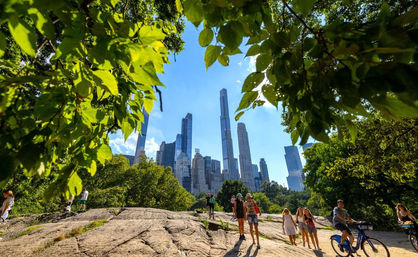 Sunny view of the Manhattan skyline framed by a leafy canopy, with people strolling and biking across a sunlit rock outcrop in Central Park.