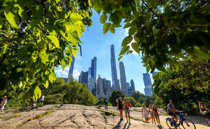 Central Park summer scene framed by leafy branches, visitors walking and a cyclist on a rocky overlook with Midtown Manhattan skyscrapers rising against a bright blue sky.