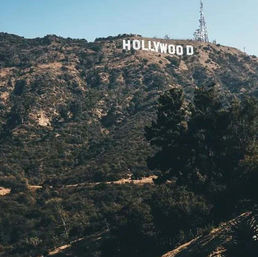 White Hollywood sign perched on sunlit Los Angeles hills, surrounded by dry chaparral and a radio tower under a clear blue sky.
