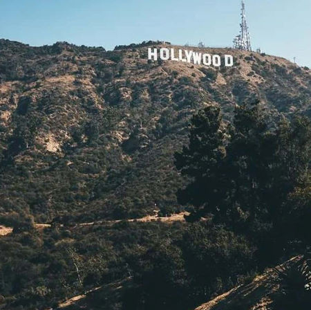 Iconic Hollywood sign perched on a sunlit Los Angeles hillside with dry chaparral, trees and a clear blue sky