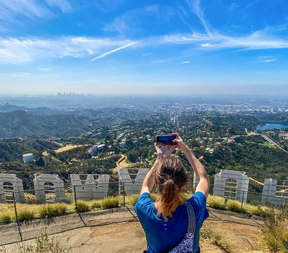 Visitor snapping a phone photo from behind giant white hillside letters, panoramic Los Angeles skyline, Hollywood Hills, winding trails and a blue reservoir under a bright blue sky.