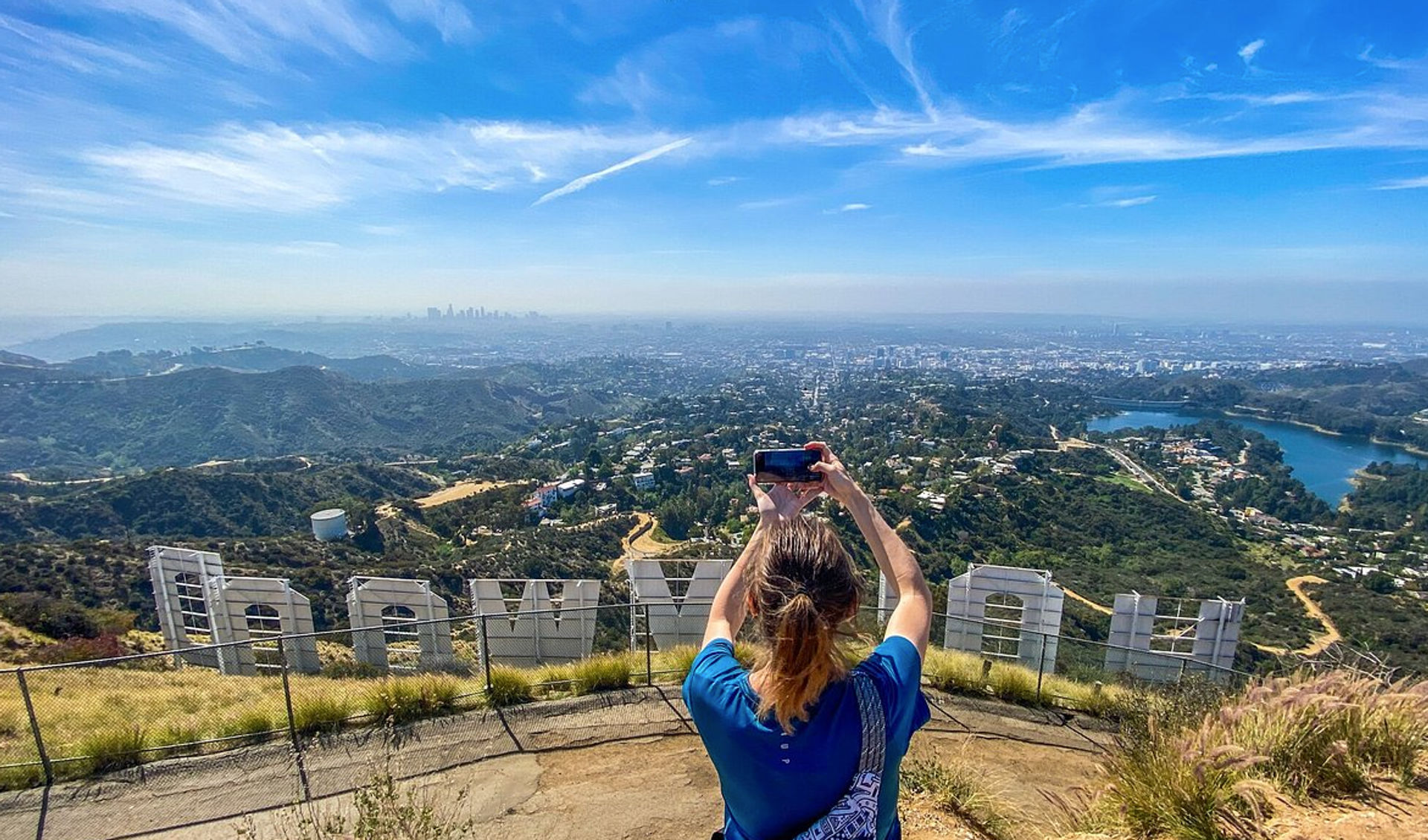 Visitor snapping a phone photo from behind giant white hillside letters, panoramic Los Angeles skyline, Hollywood Hills, winding trails and a blue reservoir under a bright blue sky.