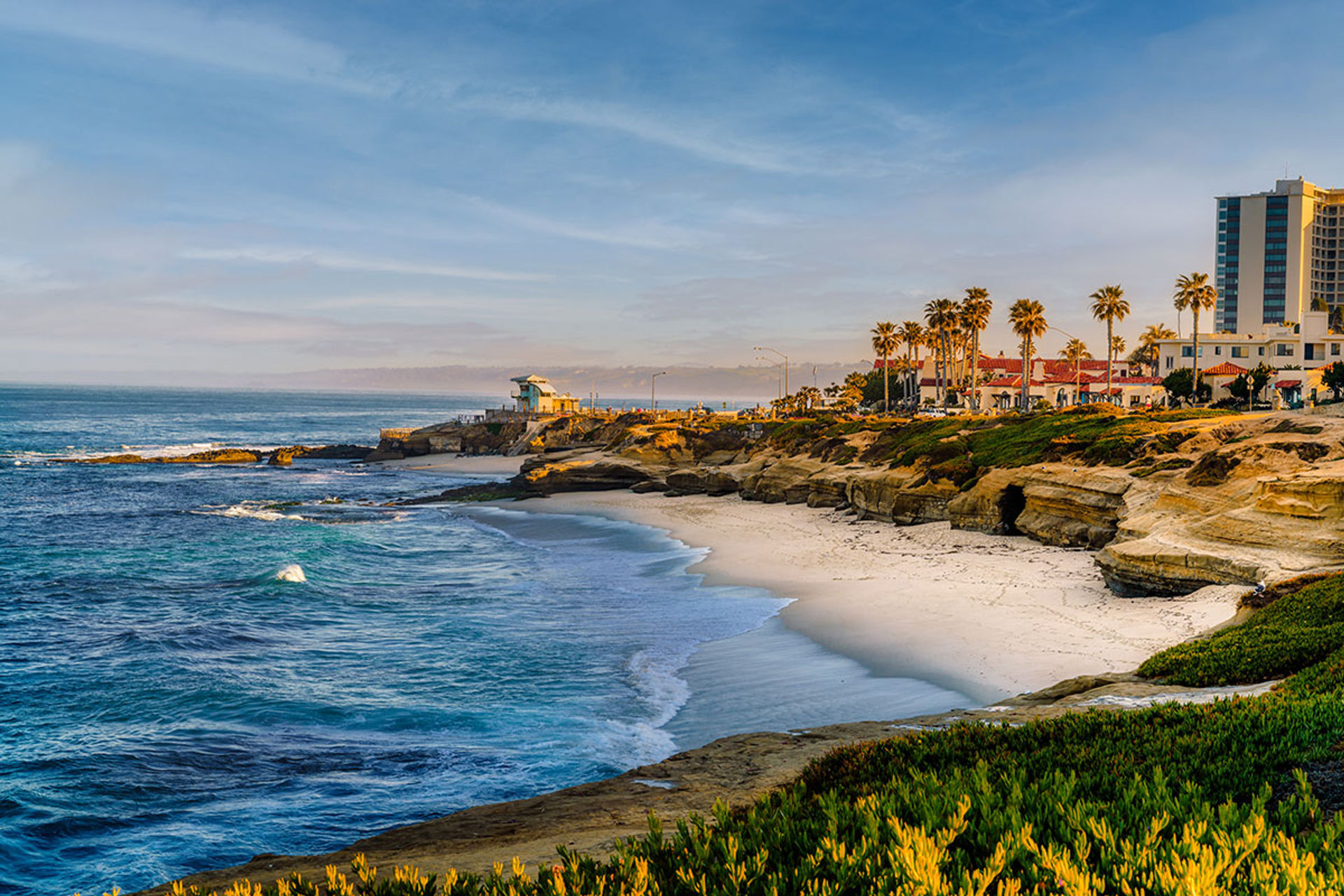 Golden-hour coastal scene: turquoise ocean waves lapping a sandy crescent beach beneath sun‑kissed sandstone cliffs lined with palm trees and seaside buildings.