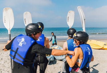 Group of kayakers in blue life jackets and black helmets laughing and grabbing paddles on a sunny sandy beach, with colorful kayaks and calm ocean waves in the background