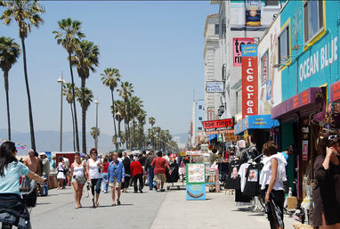 Busy Venice Beach boardwalk in Los Angeles with tall palm trees, colorful shops, street vendors and crowds on a sunny day