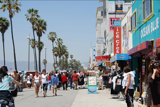 Busy Venice Beach boardwalk in Los Angeles with tall palm trees, colorful shops, street vendors and crowds on a sunny day